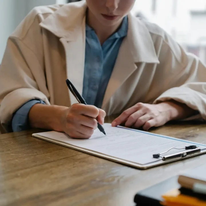 A woman in a formal setting fills out paperwork on a clipboard at an office desk.